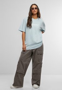 Gaiši zils pārspīlēts t-krekls ar baltu tekstu, apvienots ar brīviem pelēkiem kravas šortiem, kas aprīkoti ar vairākiem kabatām, un smilšu krāsas sporta apaviem.