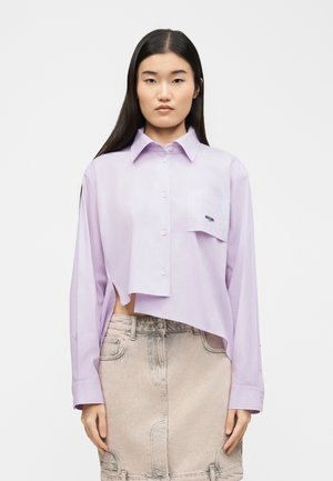 Sieviete ar gariem tumšiem matiem, valkājot violetu kroplētu kreklu ar pogām un gaiši bēšu džinsa svārkus, stāv pret vienkāršu fonu.