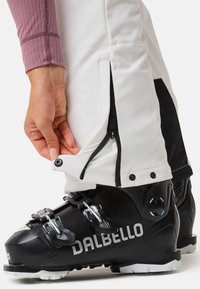 Zwarte skiboot met zilveren hardware, voorzien van verstelbare gespen. In de buurt bevinden zich witte skibroeken met rits- en drukknoopsluitingen aan de enkels.