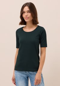 Tmavě zelené tričko s krátkým rukávem a kulatým výstřihem, vyrobené z měkké tkaniny. Má jednoduchý design, nošené s světlými modrými džíny.