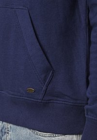 Navyblaues Baumwoll-Hoodie mit Kängurutasche, gerippten Bündchen und Saum, mit einem kleinen metallischen Logo-Akzent in der Nähe der Tasche.