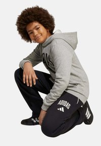Jeune personne aux cheveux bouclés portant un sweat à capuche gris Adidas et un pantalon de survêtement noir Adidas, agenouillée et souriante sur un fond uni.