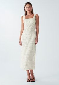 Svijetlosmeđa, bezrukavna midi haljina sa kvadratnim izrezom, ukrašena crnim točkicama. Glatka tekstura, minimalistički dizajn, bočni prorez na donjem rubu.