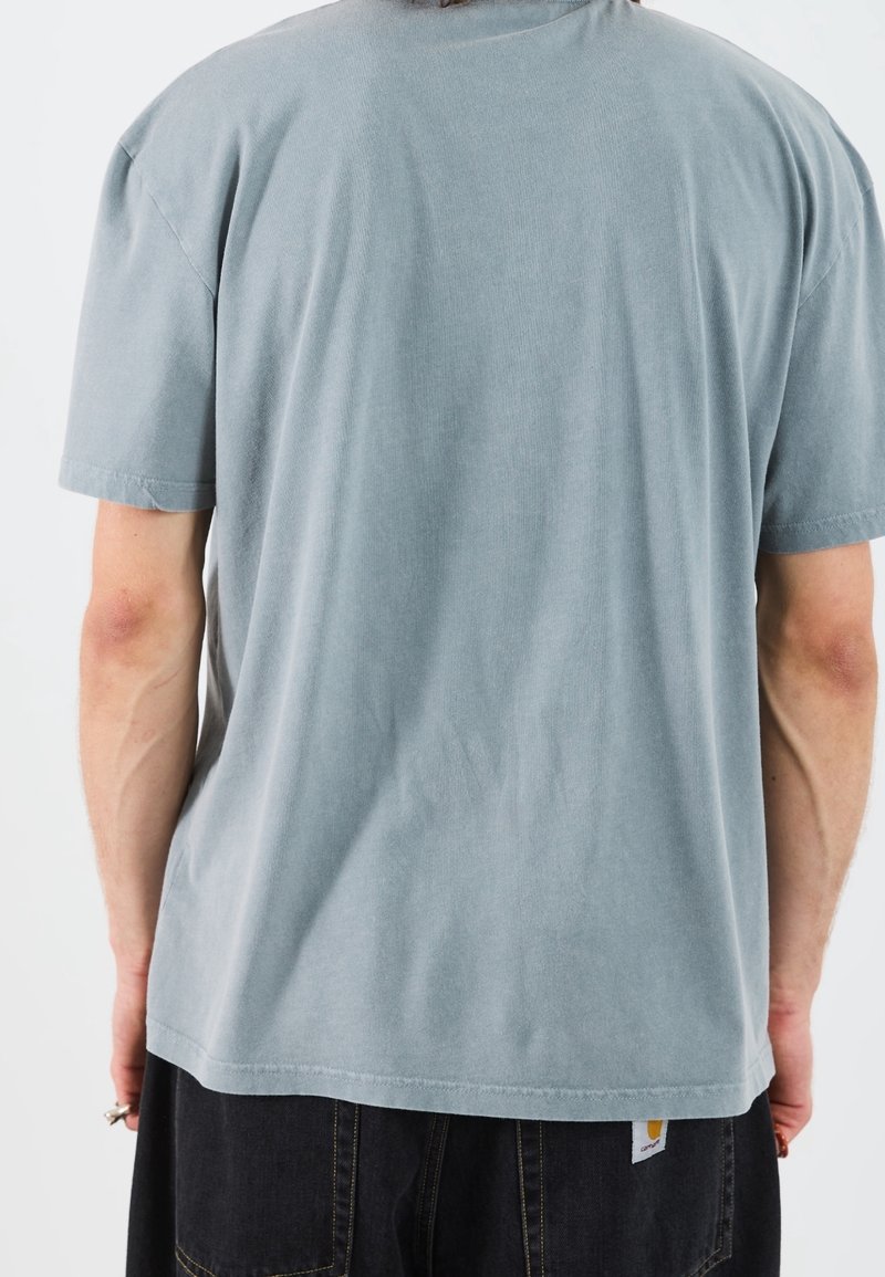 Personne portant une chemise bleu clair unie à manches courtes et un pantalon foncé vue de dos sur un fond uni.