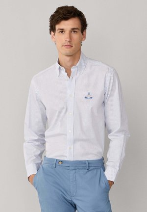 Homme portant une chemise blanche à boutons avec un motif à carreaux bleu clair et un pantalon bleu, les mains dans les poches, sur un fond gris uni.