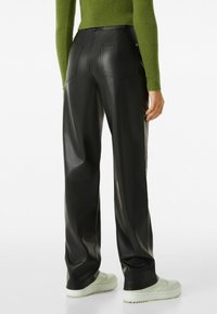 Pantaloni neri in faux pelle con una vestibilità rilassata, caratterizzati da due tasche posteriori, abbinati a sneaker chiari e a un maglione verde testurizzato.