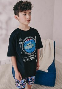 T-shirt noir avec un graphique de ballon de soccer coloré, associé à un short à motifs multicolores. L'arrière-plan inclut une chaise bleue et une couverture.