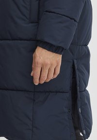Chaqueta de plumón azul marino con secciones cosidas, presenta un cierre de cremallera y botones a presión. Primer plano de una mano sosteniendo el costado de la chaqueta.