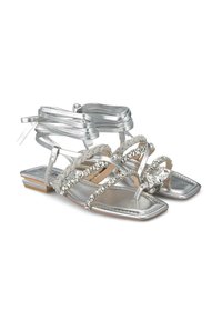 Sandales plates en argent avec un bout carré, dotées de brides métalliques, d'accents de bijoux et de rubans décoratifs enroulés autour de la cheville.