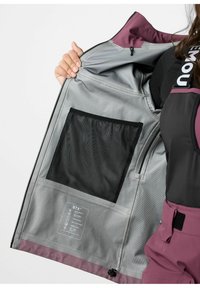 Intérieur d'une veste imperméable en gris avec une poche en maille, des accents violets, et une étiquette d'entretien détaillant les propriétés du tissu et les instructions de maintenance.