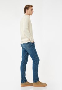 Suéter color beige claro con cuello redondo, combinado con jeans de mezclilla azules. Zapatos color marrón completan el look, con una textura suave y un ajuste casual.
