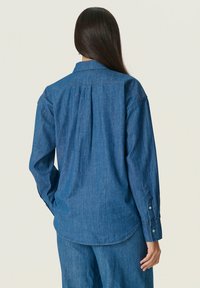 Cămașă din denim albastru închis cu mânecă lungă, cu guler clasic, detalii cu pliere pe spate și mansete cu nasturi. Textură moale cu un finisaj uniform.