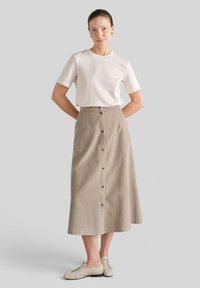 Jasno-beżowa spódnica do kolan o pionowej teksturze prążkowanej, z guzikami z przodu, w połączeniu z gładką białą koszulką z krótkim rękawem i jasnymi butami.