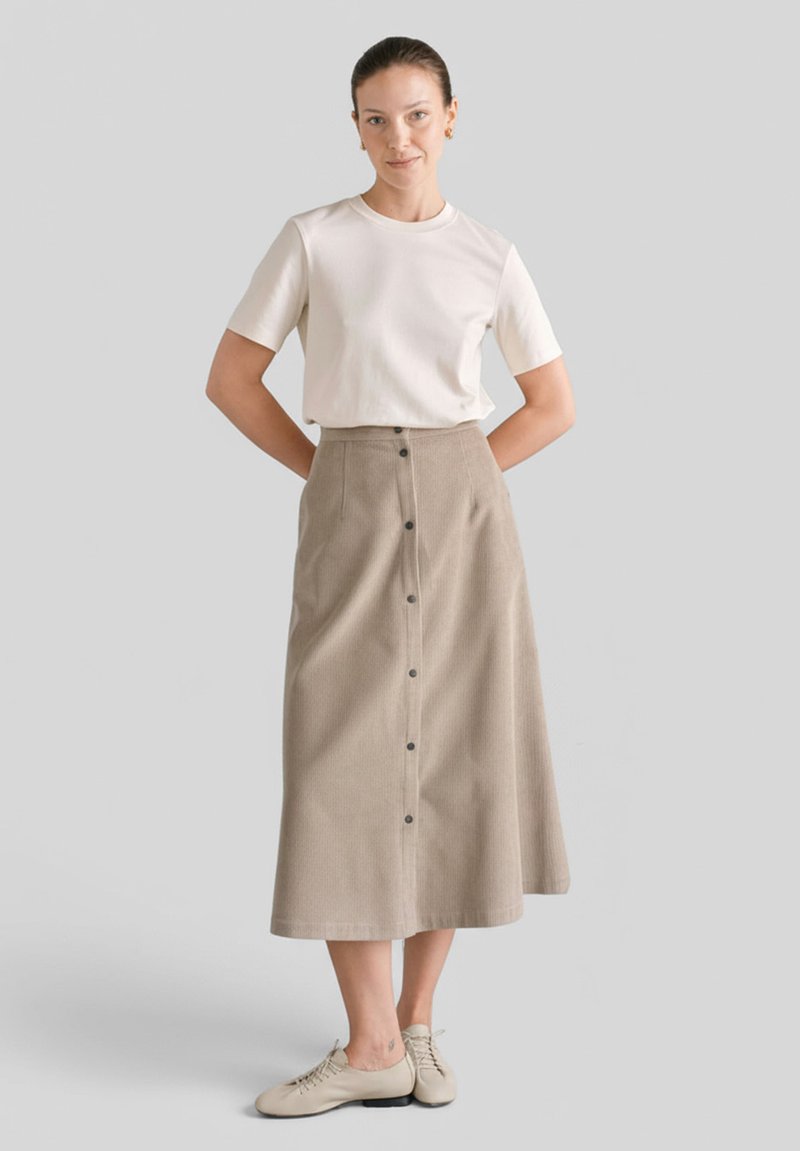 Jasno-beżowa spódnica do kolan o pionowej teksturze prążkowanej, z guzikami z przodu, w połączeniu z gładką białą koszulką z krótkim rękawem i jasnymi butami.