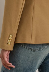 Beige teksturert blazer med gullknappdetaljer på ermet, kombinert med lyseblå denimjeans med standard baklomme.