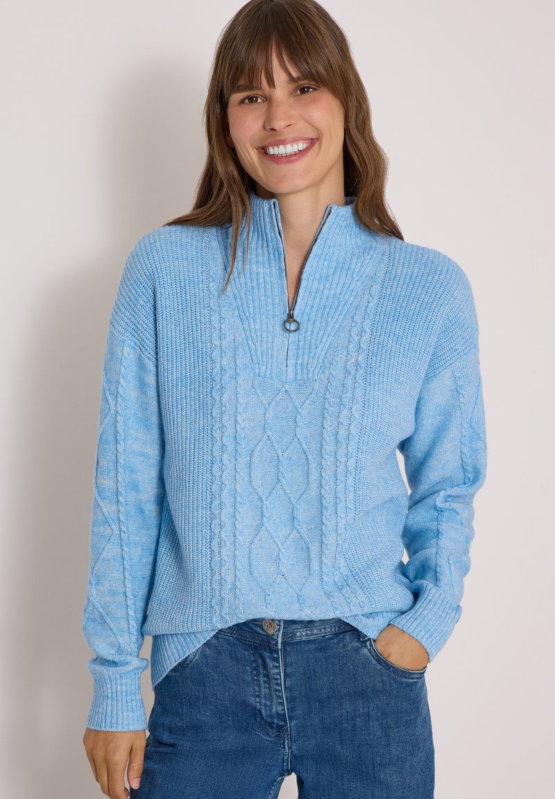 Femme souriante portant un pull torsadé bleu clair avec une fermeture éclair à l'avant et un jean bleu, posant avec une main dans la poche sur un fond blanc.