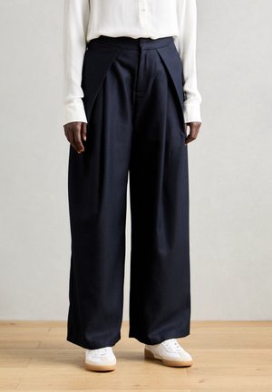 Pantaloni a gamba larga blu scuro con fronte plissettato, realizzati in tessuto morbido; abbinati a una camicia bianca con bottoni e sneakers bianche.