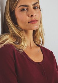 Burgundy cardigan with a scoop neckline, button closure, and soft texture. Features light sheen and delicate tattoo visible on the collarbone.