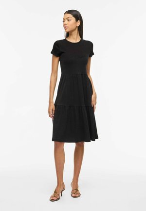 Femme portant une robe midi noire à manches courtes avec une jupe à volants, associée à des escarpins dorés à bride en T, posant devant un fond blanc.