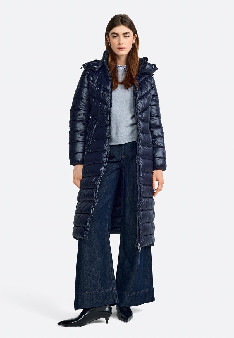 Giacca più leggera blu navy con zip frontale e cappuccio, caratterizzata da un design a trapunta e tasche laterali; abbinata a pantaloni in denim scuro a gamba larga.