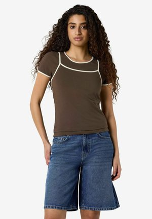 Ung kvinde med langt krøllet hår iført en brun kortærmet top med hvide kanter og blå cowboyshorts til knæet.