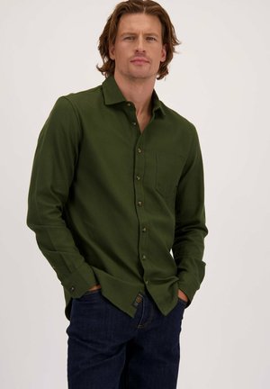 Man met middelmatig lang haar, gekleed in een donkergroen knoopoverhemd en donkerblauwe spijkerbroek, met handen gedeeltelijk in zijn zakken, op een neutrale achtergrond.