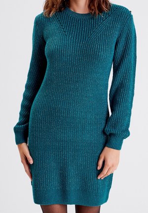 Femme portant une robe-pull à manches longues en maille côtelée couleur sarcelle avec un léger éclat, debout devant un fond clair uni.