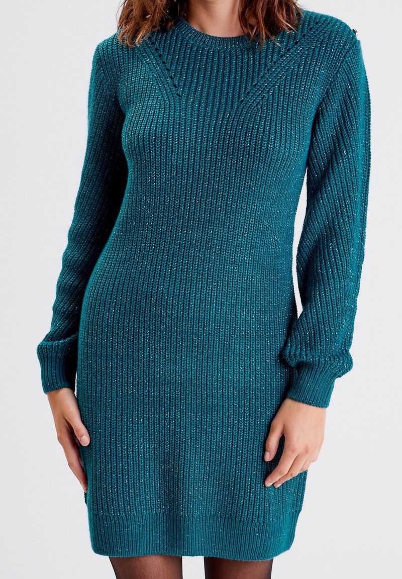 Femme portant une robe-pull à manches longues en maille côtelée couleur sarcelle avec un léger éclat, debout devant un fond clair uni.