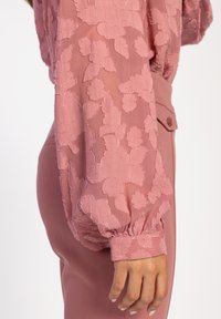 Langärmlige, durchsichtige rosa Bluse mit strukturierten Blumenstickereien. Der Ärmelaufschlag ist gerafft und hat eine Knopfdetails, kombiniert mit maßgeschneiderten rosa Hosen.