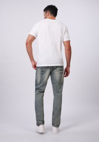 Weißes T-Shirt mit kurzen Ärmeln, hellblaue Jeans mit Ausbleichungen und weiße Sneaker. Von hinten vor einem schlichten Hintergrund gesehen.