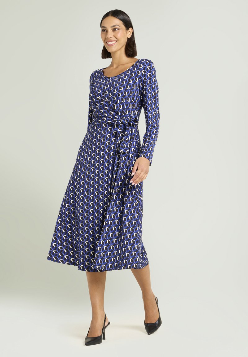 Niebieska sukienka na długi rękaw z geometrycznym wzorem, z talią podkreśloną paskiem, zwiewną spódnicą, zestawiona z czarnymi szpilkami. Gładka faktura tkaniny.