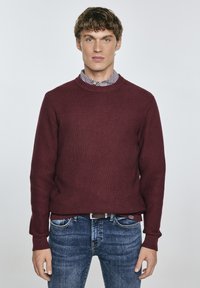 Burgunderfarbener Strickpullover mit geripptem Muster und Rundhalsausschnitt, über einem karierten Hemd getragen, kombiniert mit blauen Jeans.