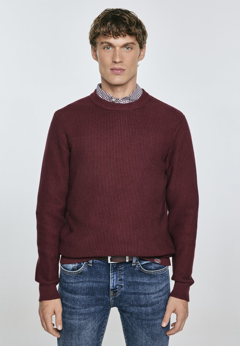 Burgunderfarbener Strickpullover mit geripptem Muster und Rundhalsausschnitt, über einem karierten Hemd getragen, kombiniert mit blauen Jeans.