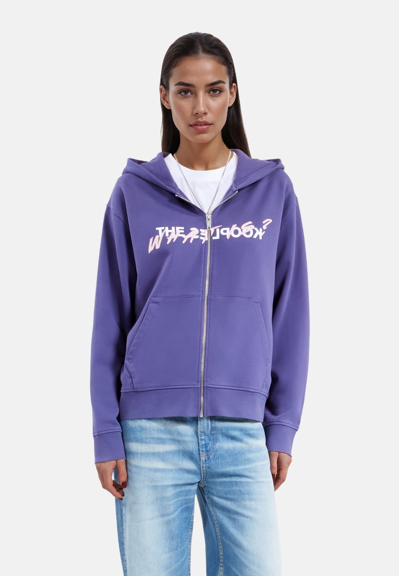 Felpa con zip viola, dotata di tasca frontale e cappuccio, con testo grafico "WHAT'S GOOD?" in bianco. Il materiale sembra morbido e presenta una vestibilità casual.