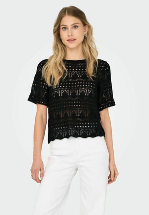 Sort hæklede top med korte ærmer, der har et åbent stingmønster og bølget kant. Modellen står med hvidt bukser mod en neutral baggrund.
