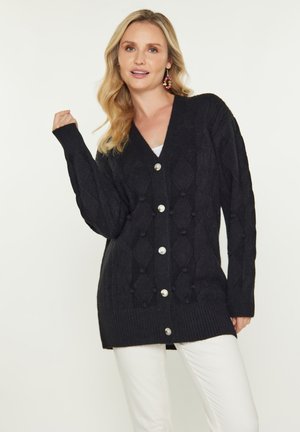 Cardigan nero in maglia con motivo a rombi, vestibilità oversize, bottoni argentati e polsini a costine. Indossato sopra una maglietta bianca e abbinato a pantaloni bianchi.