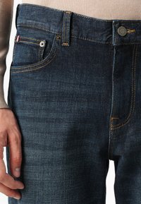 Zoom na temno modre kavbojke z bež šivi, sprednjim žepom in roko, počivajočo ob stegnu, oblečeno s svetlo bež majico.