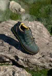 Zelená a žlutá turistická bota s hladkým svrškem, texturovanou špičkou a zpevněnou patou. Obsahuje kontrastní šňůrky a švy.