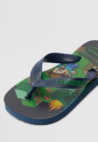 Zila gumijas čīpa ar pikselētu Minecraft dizainu. Iekļauj teksturētu siksnu, zaļu "havaianas" logo un rakstainu zolīti.