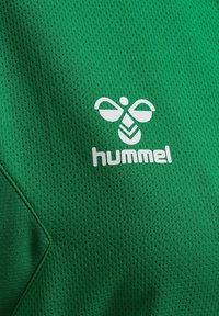 Logo d'abeille stylisé blanc avec le texte « hummel » sur tissu vert texturé, probablement un maillot de sport ou un vêtement de sport.