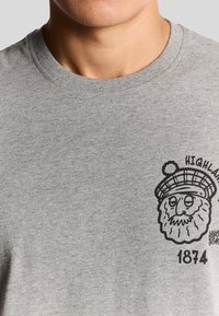 Grijze katoenen T-shirt met een zwarte grafiek van een leeuw in een baret en de tekst "HIGHLAND 1874" op de linkerborst. Ronde hals stijl.