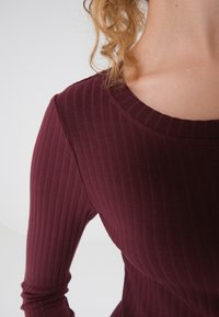 Lähikuva henkilöstä, joka on pukeutunut ribbimäiseen, pitkähihaiseen viininpunaisiin yläosaan ja jonka kiharat hiukset näkyvät olkapäällä.