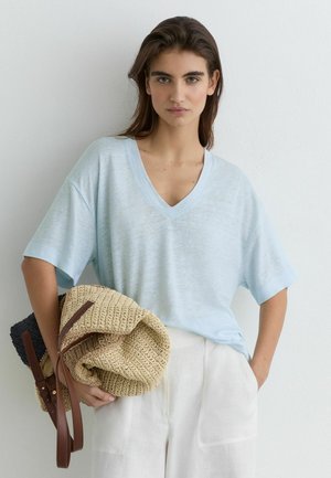 Kvinde med langt brunt hår iført lyseblå V-hals T-shirt og hvide bukser, der holder to flettede tasker med brune læderstropper.