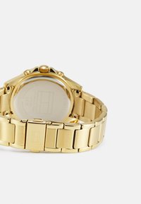 Montre en métal doré avec une finition polie, dotée d'un boîtier rond et d'un bracelet à maillons. Le dos présente un verre transparent et des détails gravés.