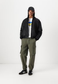 Schwarze Bomberjacke, weißes T-Shirt mit grafischem Druck und blau-gelbem Logo, olivgrüne Cargo-Hose, schwarze Kappe und schwarze Adidas-Sneaker.