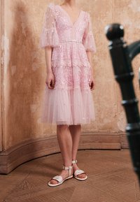Robe rose avec des manches bouffantes transparentes, broderies florales et accents en dentelle. La jupe a des superpositions de tulle, portée avec des sandales blanches ornées de nœuds.