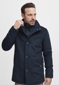 Mornarsko plavi vuneni kaput s visokim ovratnikom, kopčanjem na dugmad i bočnim džepovima. Sadrži podstavu od jastučića i diskretnu oznaku brenda blizu rubova.