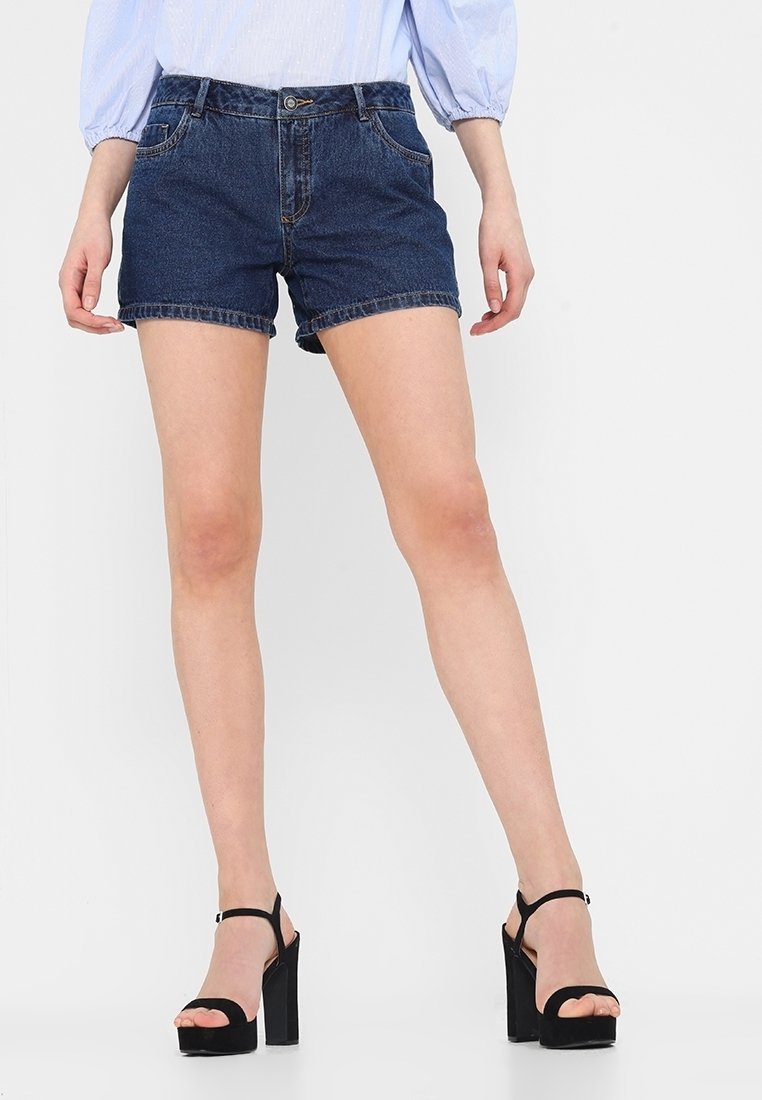 Tummansiniset farkkushortsit klassisella istuvuudella, viisi-tasku-mallilla ja käärityillä lahkeilla, yhdistettynä mustiin korokepantseihin.