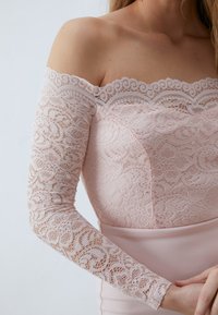 Robe décolletée en rose clair, avec des manches en dentelle délicates et un corsage lisse ajusté. Bords festonnés délicats et motifs floraux.
