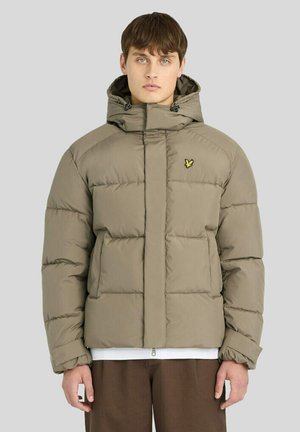 Young man wearing a beige padded jacket with a hood and a small yellow bird logo on the chest, standing against a plain light background.